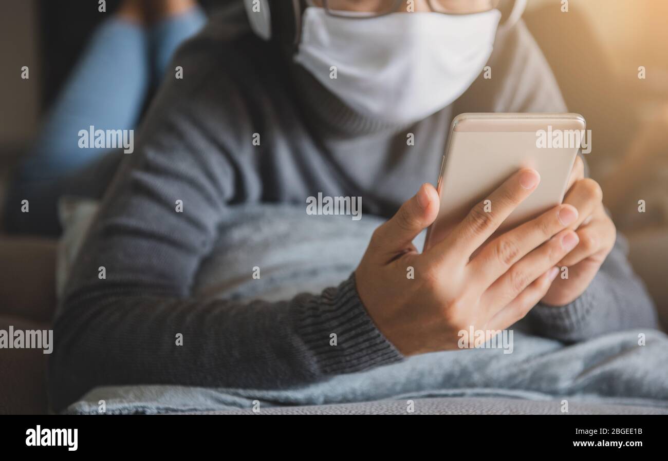 Young woman wearing mask staying at home listening music with smartphone, headphone relaxing emotion. Prevent the spread of coronavirus quarantine. Stock Photo