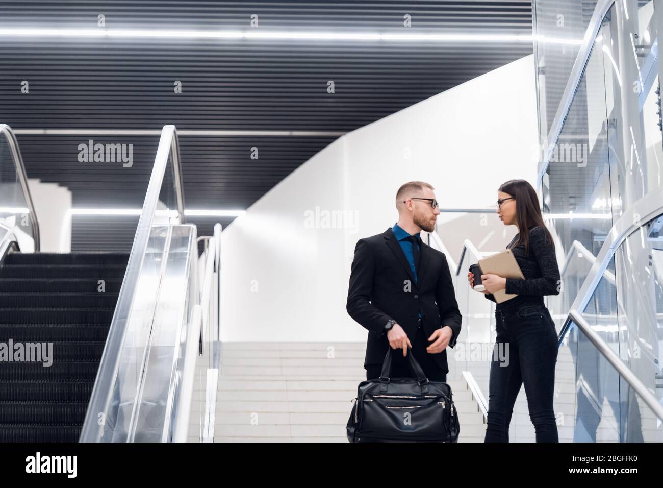 Business couple on a stairs talking and smiling. Man wearing suit and glasses with a bag talking with a woman holding tablet computer. Stock Photo