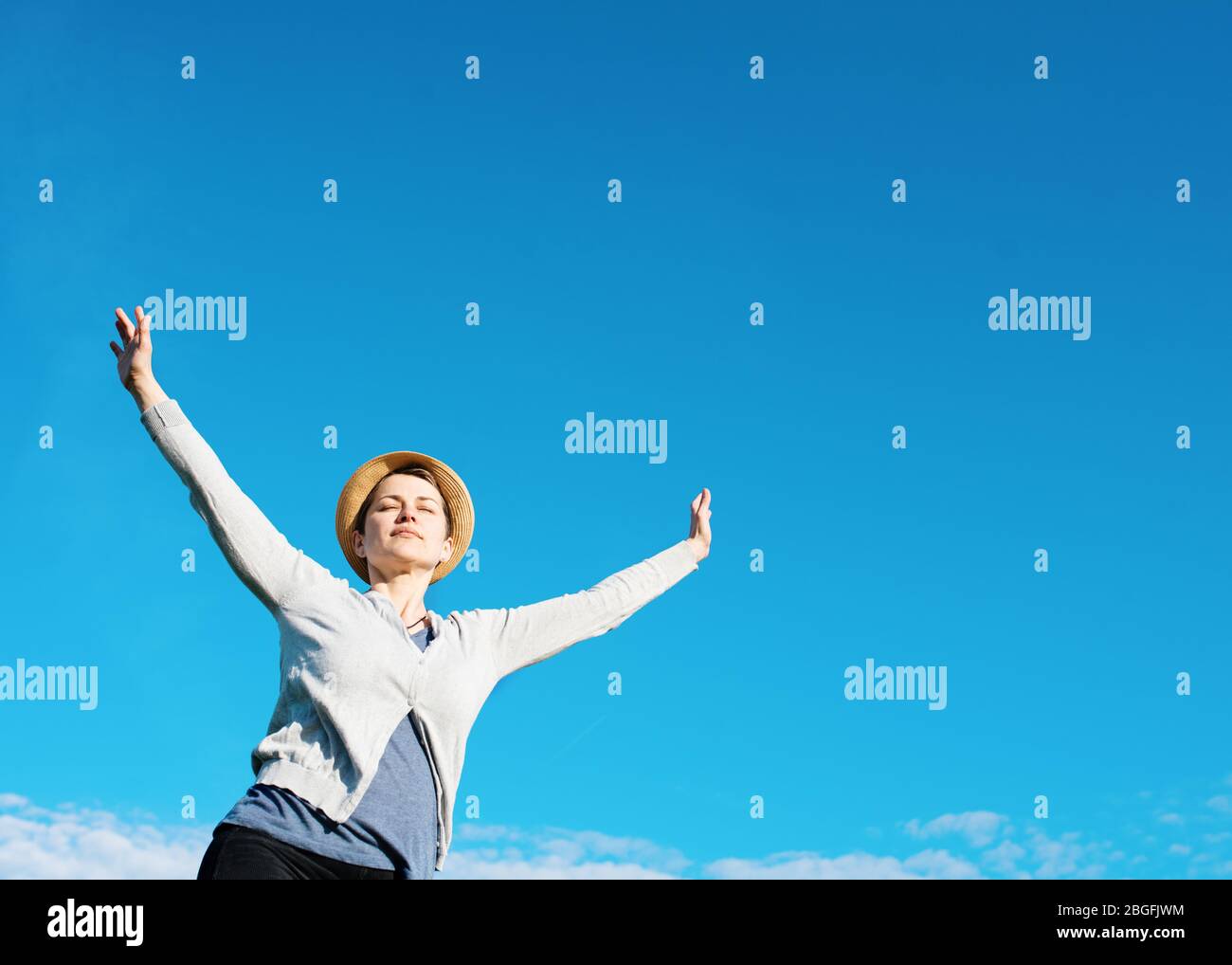 Happy woman wearing grey top, straw hat,  raised her hands high against blu sky Stock Photo