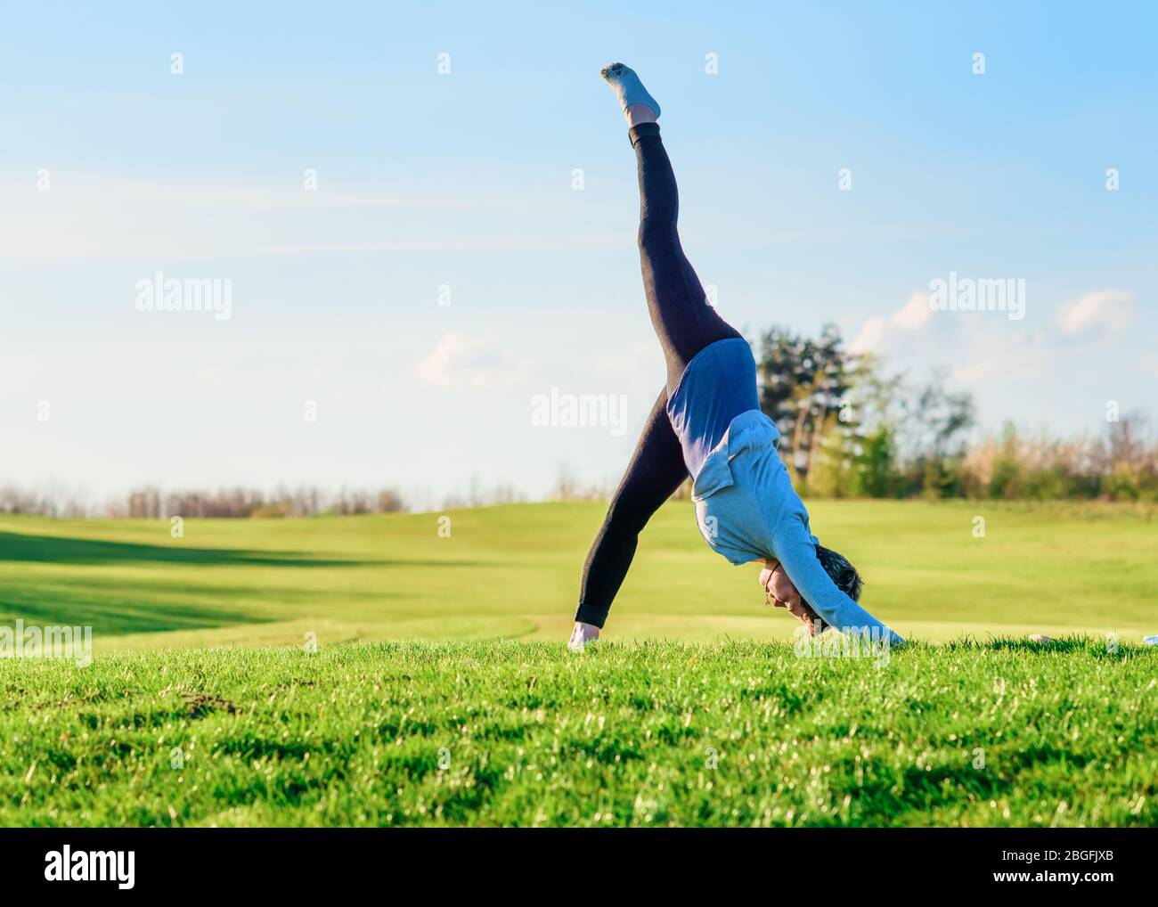 Woman wearing a grey top and black trousers doing yoga on a meadow in sunny summer day against the blue sky Stock Photo