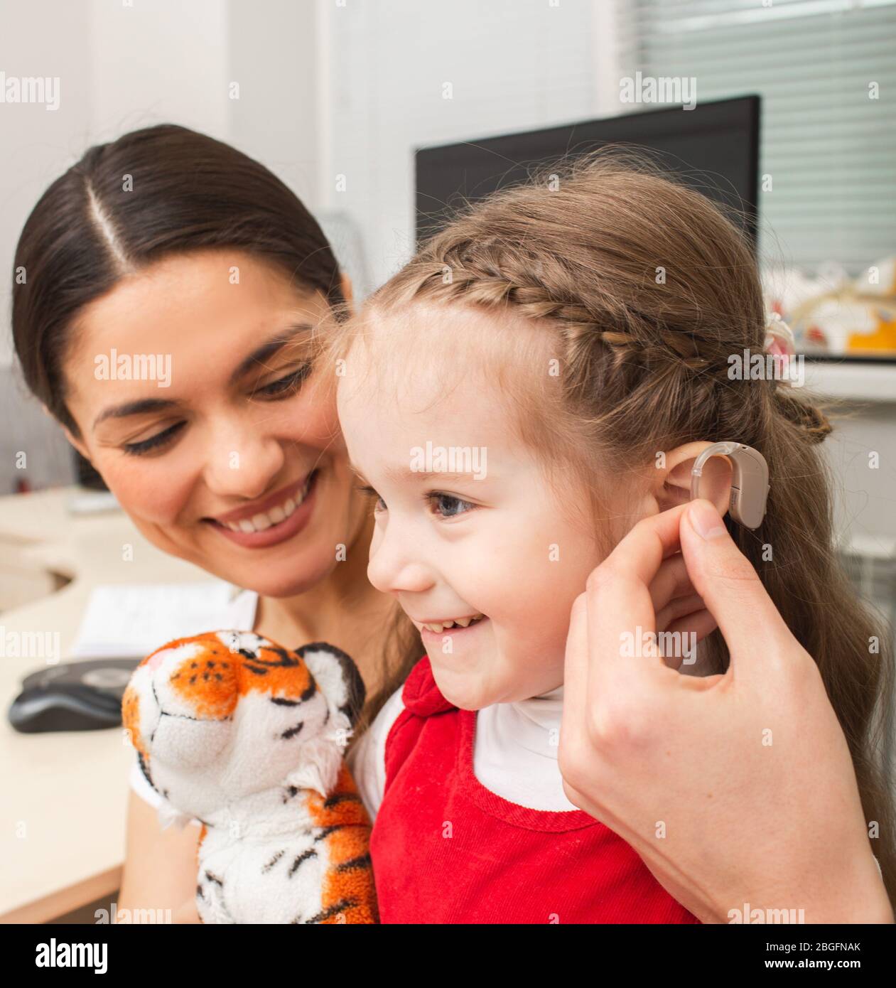 Child is not afraid that she needs to use a hearing aid on her ear. Mother of child is very pleased that her daughter will hear the world. Deafness tr Stock Photo