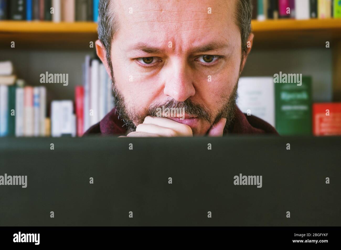 Adult man working from home office. Bearded man dedicated working online from home on computer laptop, book shelves behind him. Close up. Stock Photo