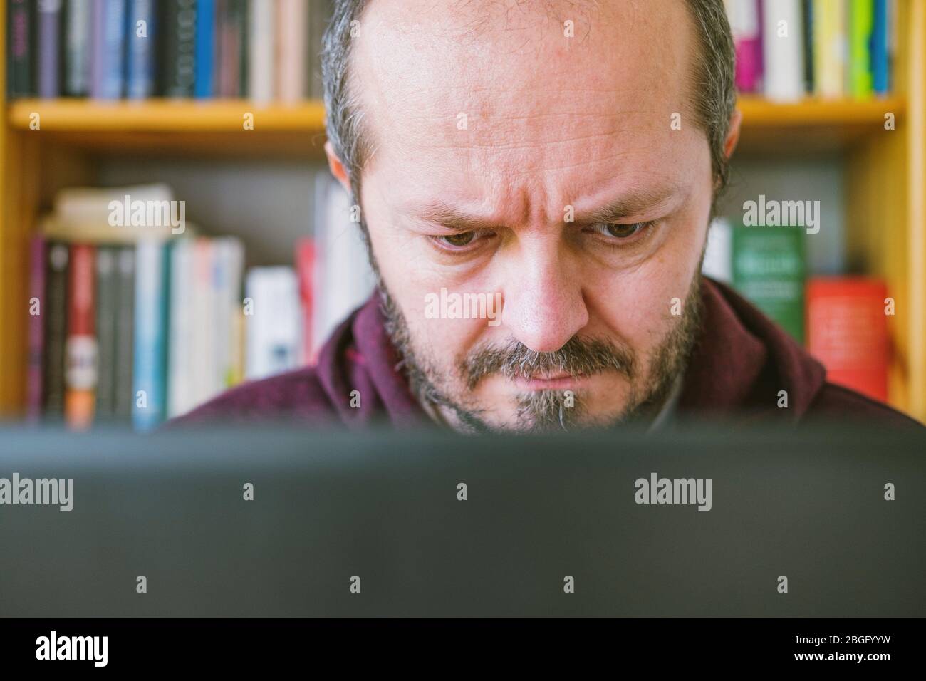 Adult man working from home office. Bearded man dedicated working online from home on computer laptop, book shelves behind him. Close up. Stock Photo