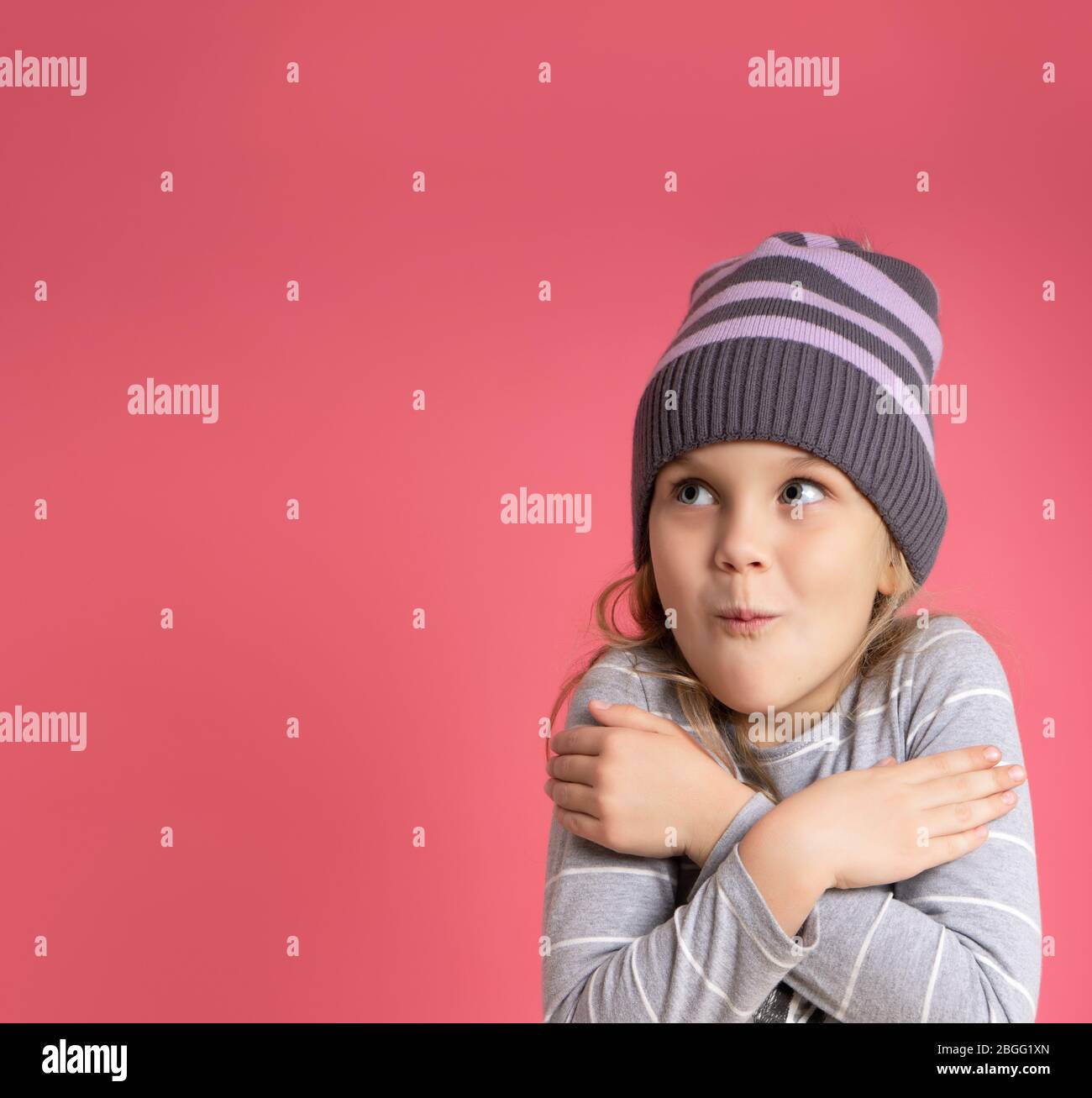 Little blonde girl in gray striped blouse and hat. She hugging herself, looking frozen, posing against pink studio background Stock Photo