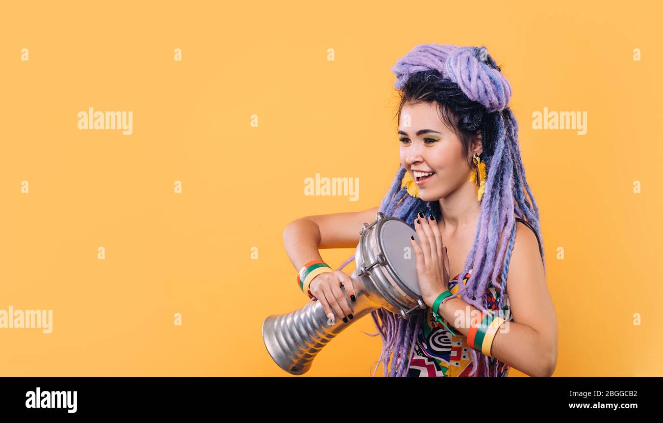 Hipster woman with violet dreadlocks and colored clothes playing on ethical mini drum on yellow background Stock Photo