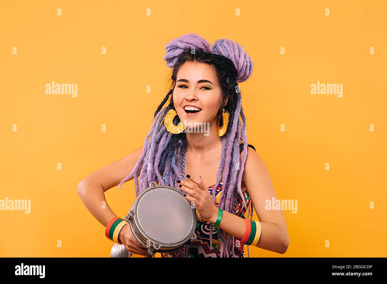 Rastafarian woman playing on ethical mini drum. Portrait woman with violet dreadlocks on a yellow background Stock Photo