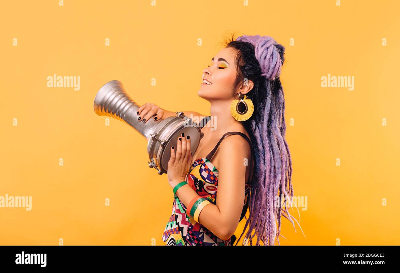Rastafarian woman with violet dreadlocks and colored clothes playing on ethical mini drum on yellow background Stock Photo