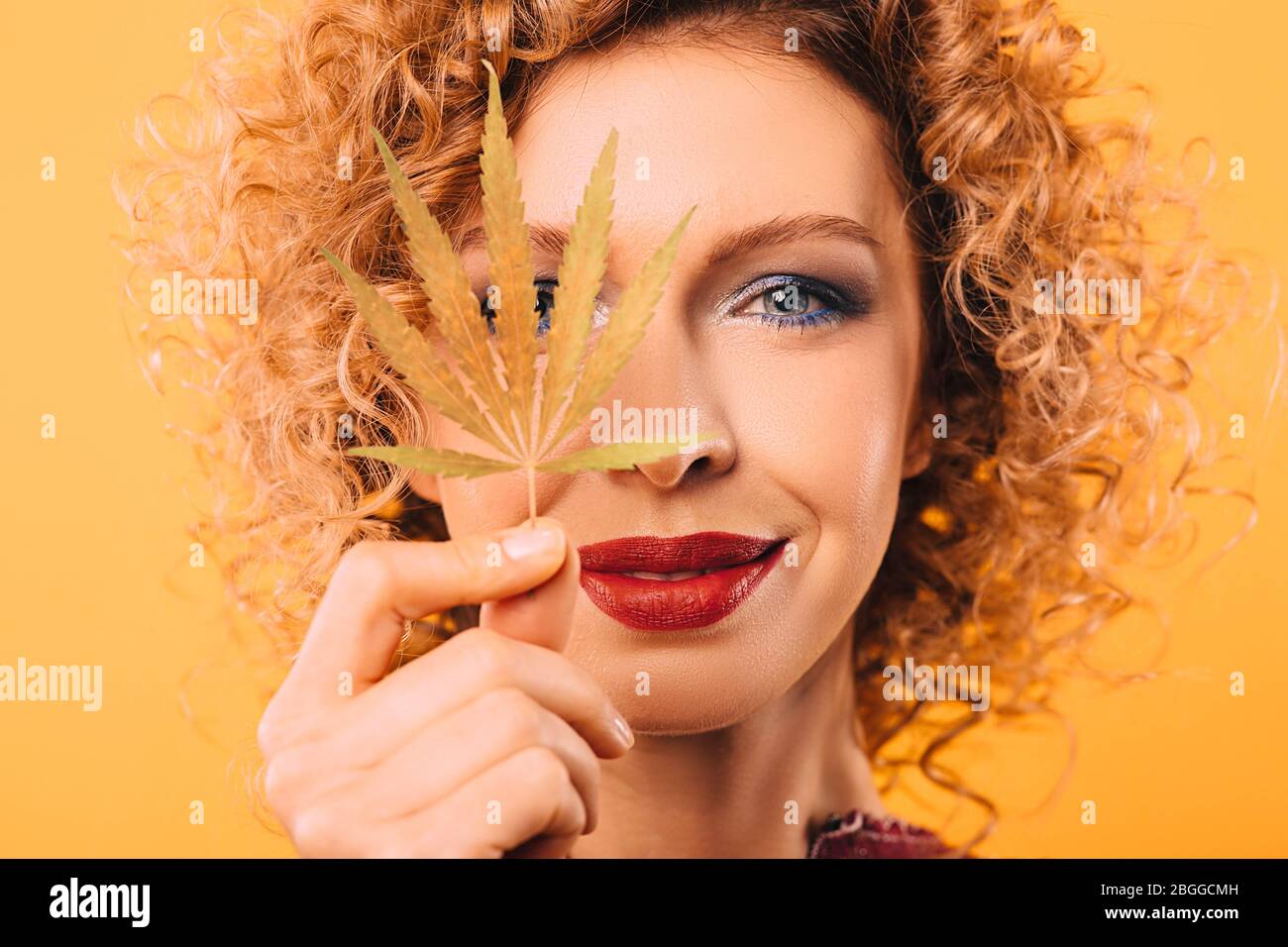 Portrait of a curly woman with a leaf of marijuana in her hand. Face close-up on a yellow background. Legalization of drugs Stock Photo