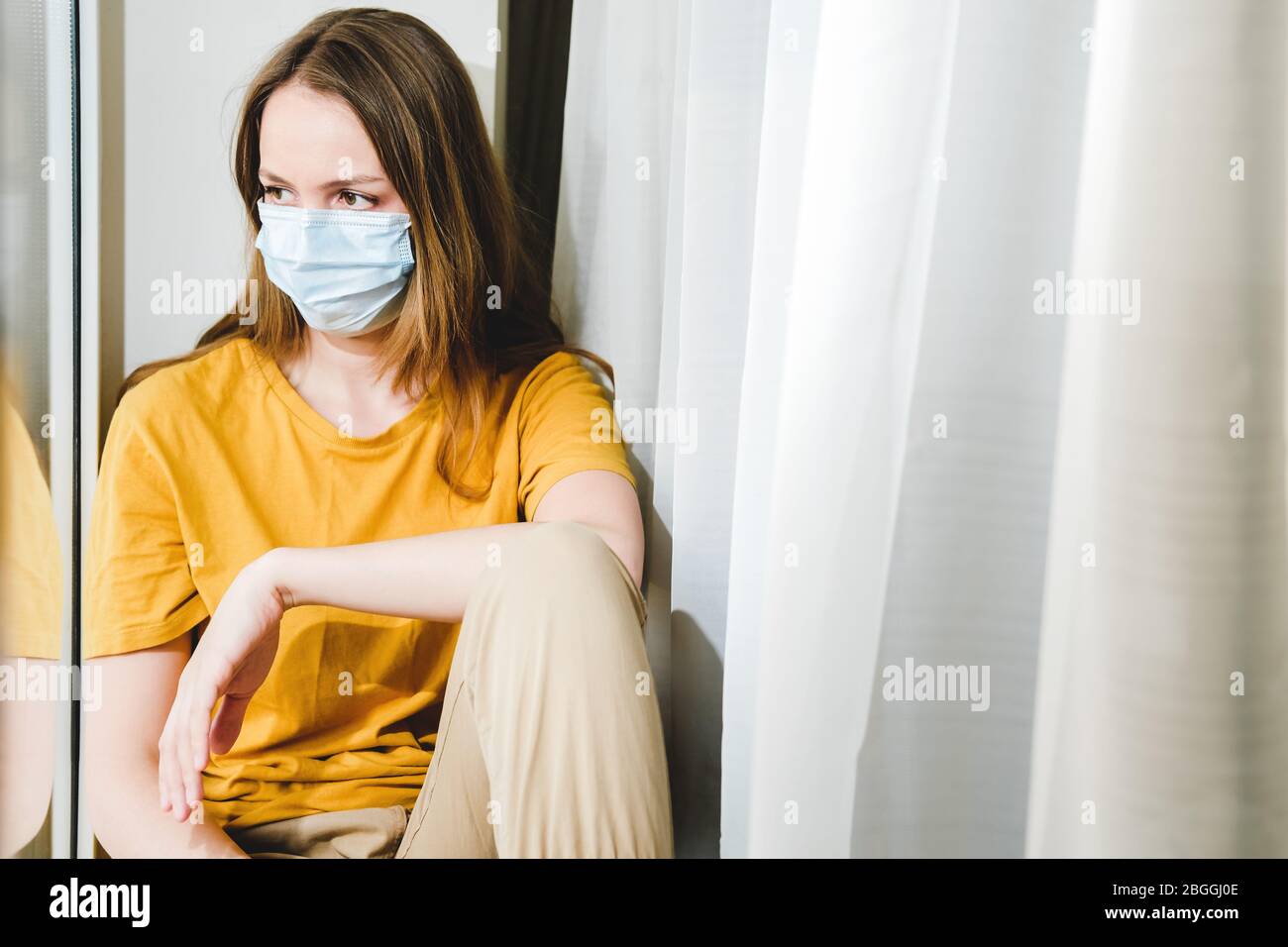 Sad girl in medical protective mask and yellow t-shirt sitting on the windowsill during a pandemic quarantine coronavirus Covid-19. Isolation at home Stock Photo