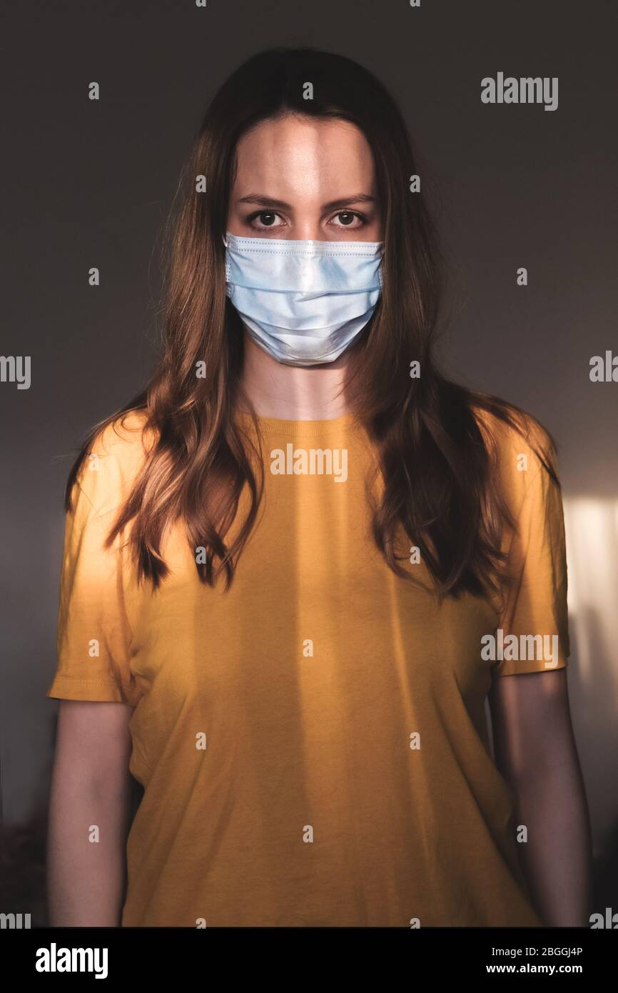 Sad girl in medical protective mask and yellow T-shirt standing at her house and looking into the camera. Covid-19 coronavirus pandemic. Isolation at Stock Photo