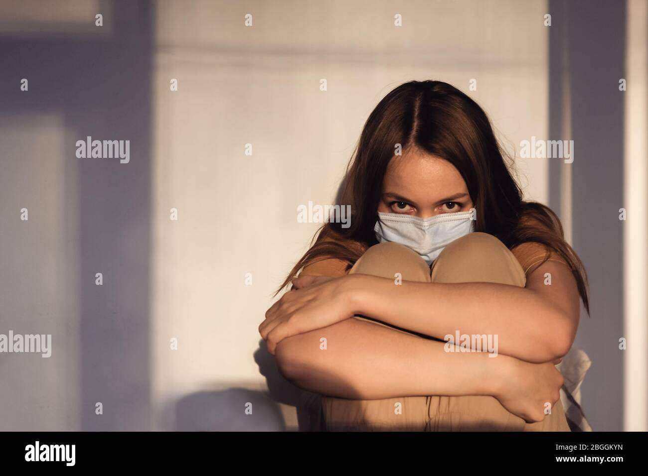 Sad girl in medical protective mask and yellow T-shirt sitting on the bed and looking into the camera. Covid-19 coronavirus pandemic. Isolation at hom Stock Photo