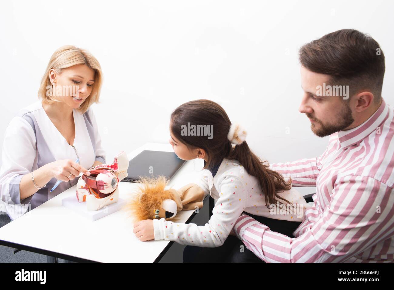 Woman optometrist shows a model of human eye to a little girl patient and her dad in ophthalmologic office. Diagnosis of vision in children Stock Photo