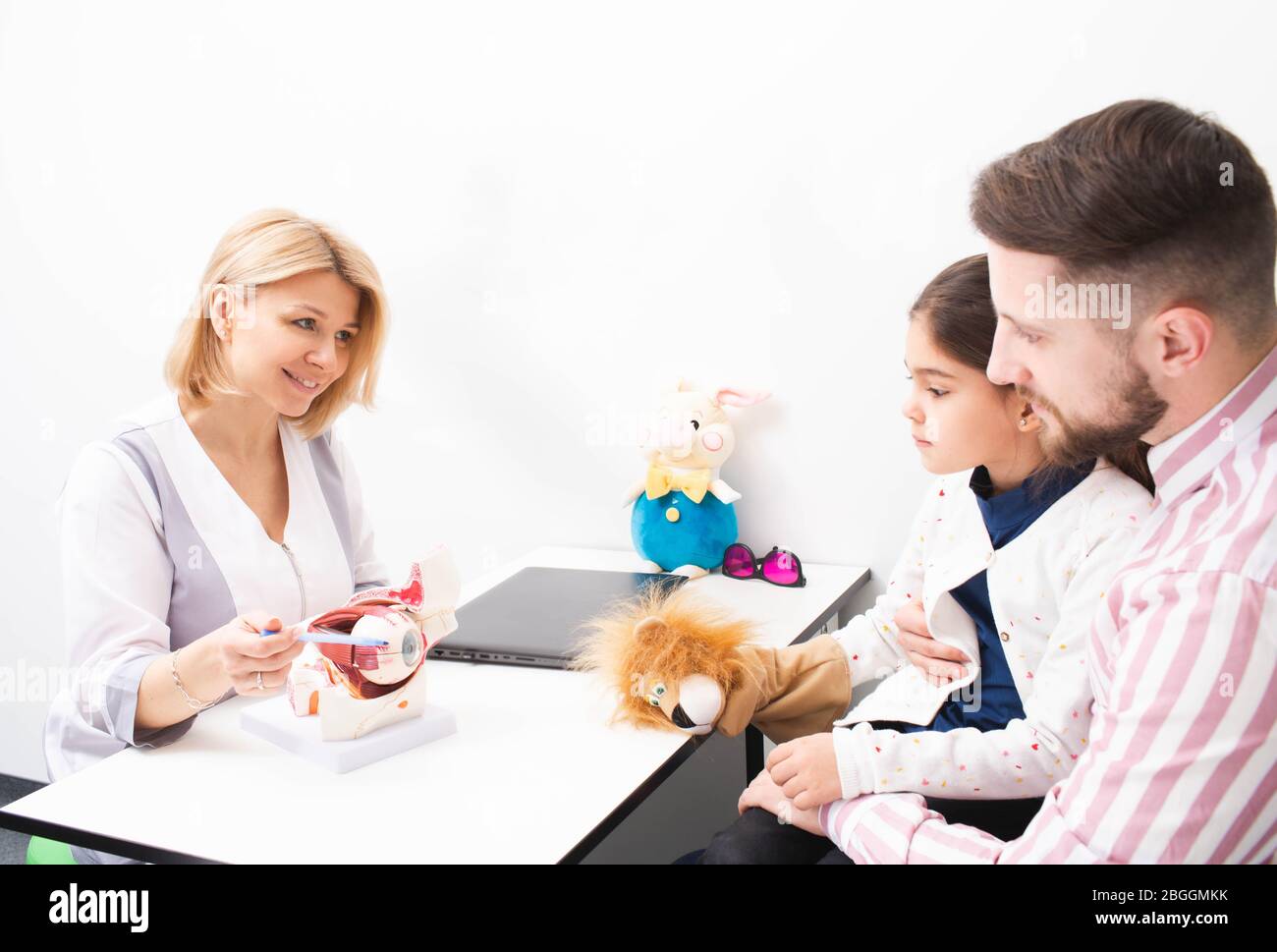 Woman optometrist shows a model of human eye to a little girl patient and her dad in ophthalmologic office. Correction of vision in children Stock Photo