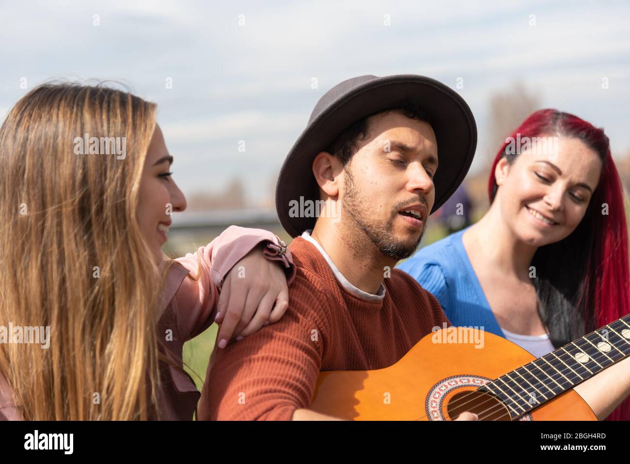 A Hispanic boy in a hat plays the guitar alongside two Caucasian girls in a city park Stock Photo