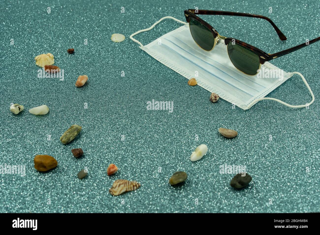 Mask protection with man sunglasses, stones and shells on ground. No holiday, stay at home. Covid-19 theme for travel. Stock Photo