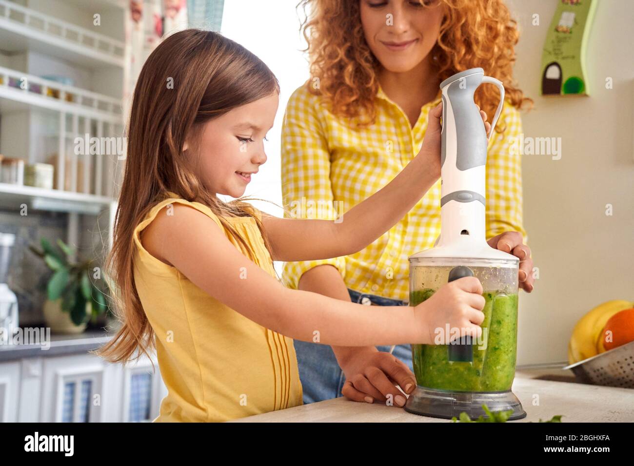 Adult mother and her little daughter making smoothie, using blender