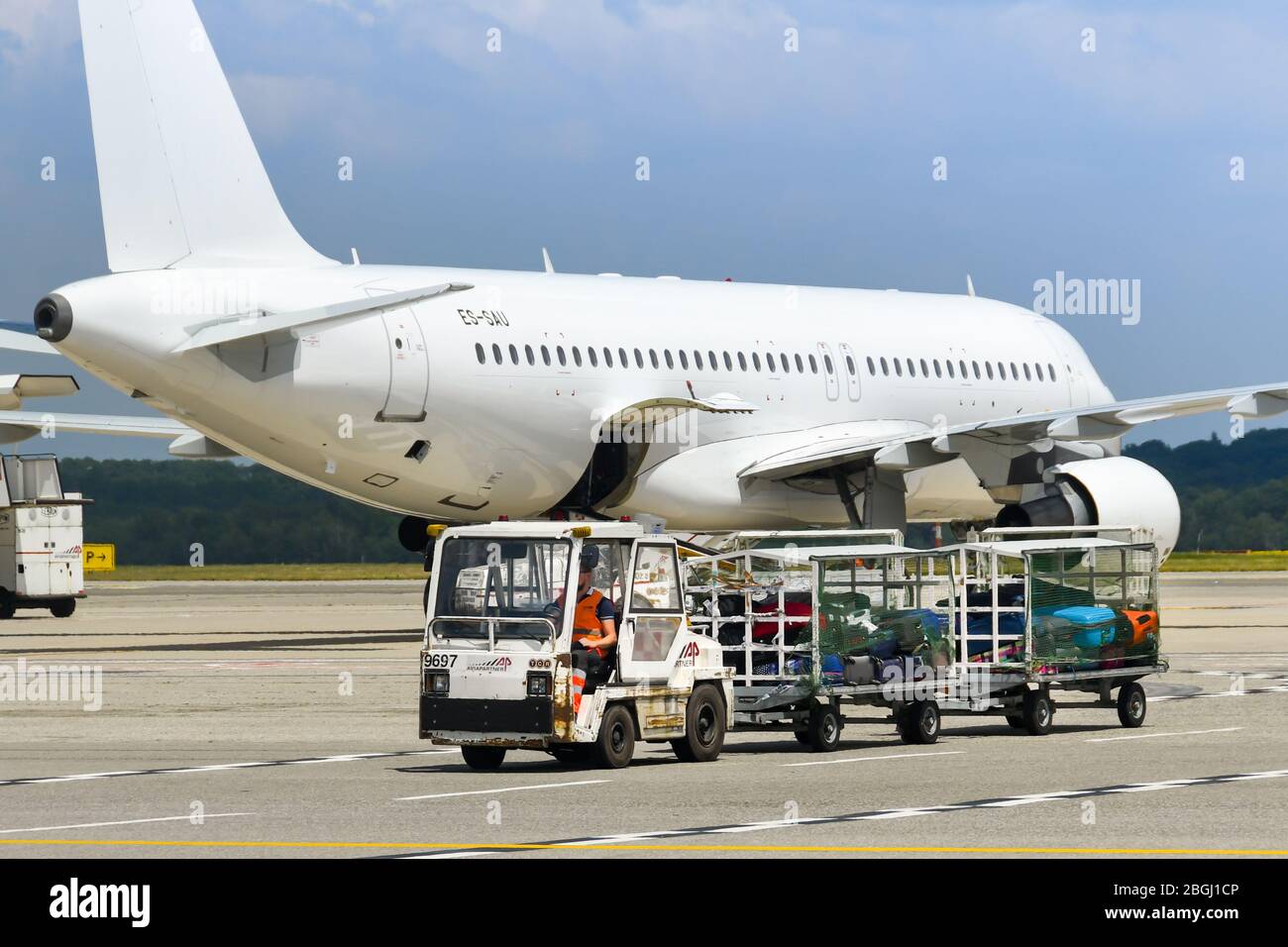 MILAN, ITALY - JUNE 2019: Small tractor pulling luggage trolleys across the apron at Milan Malpensa Airport. In the background, a plane is taxiing Stock Photo