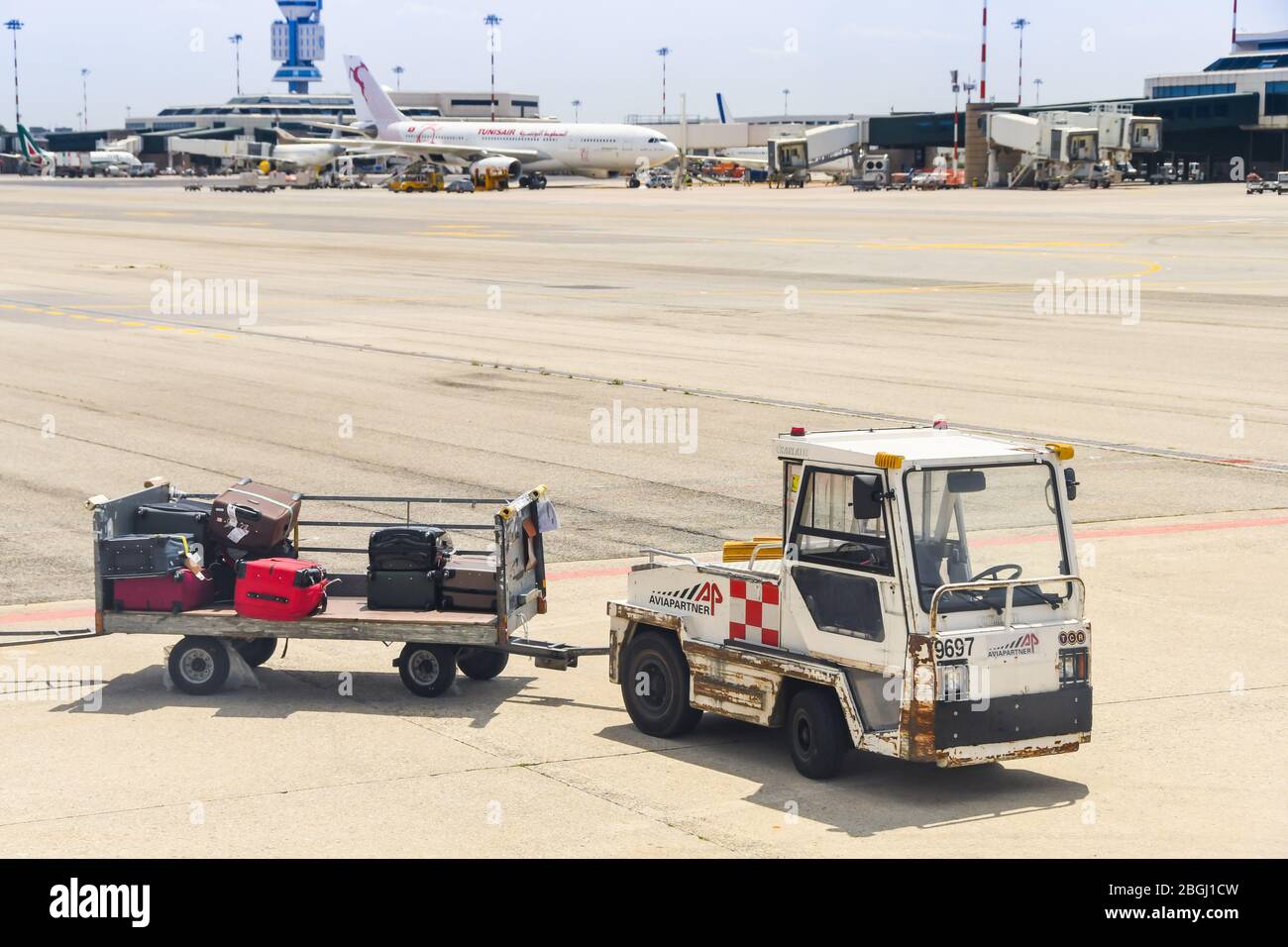 MILAN, ITALY - JUNE 2019: Small tractor with luggage trolleys parked on the apron at Milan Malpensa Airport. In the background is a plane Stock Photo