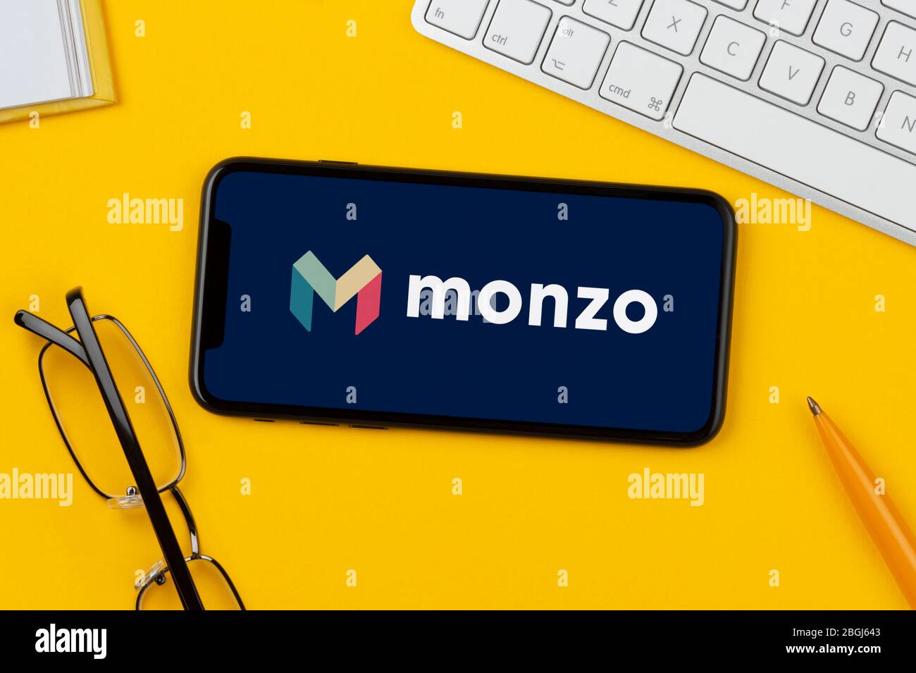 A smartphone showing the Monzo Bank logo rests on a yellow background along with a keyboard, glasses, pen and book (Editorial use only). Stock Photo