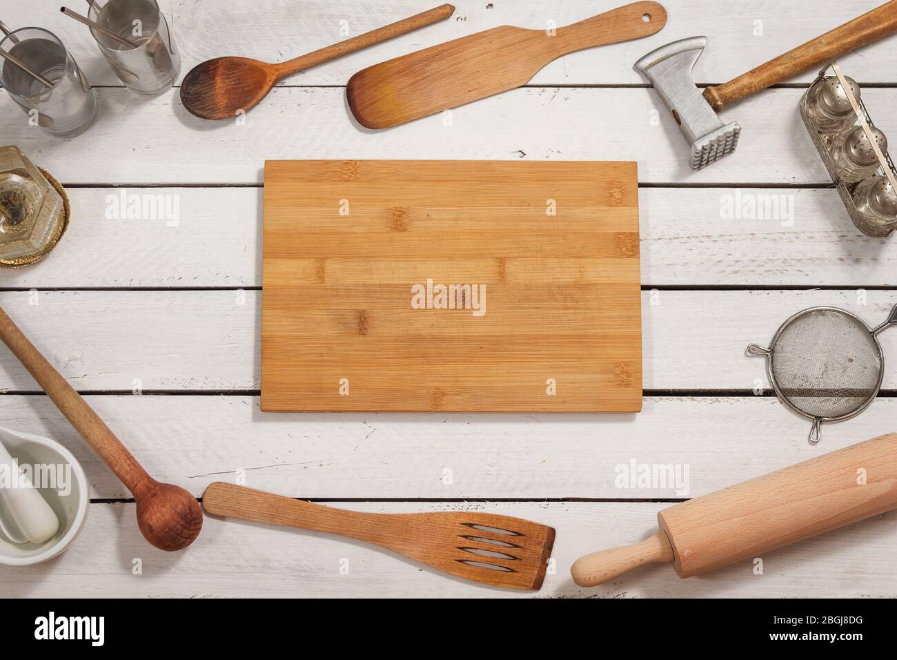 The wooden kitchen board lies on a white table. Around there are old stylish wooden kitchen utensils forming a background. Stock Photo