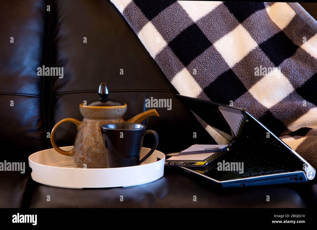 Working at home on black leather sofa with tea cup, tea pot, laptop, notebook, and comfortable blanket Stock Photo