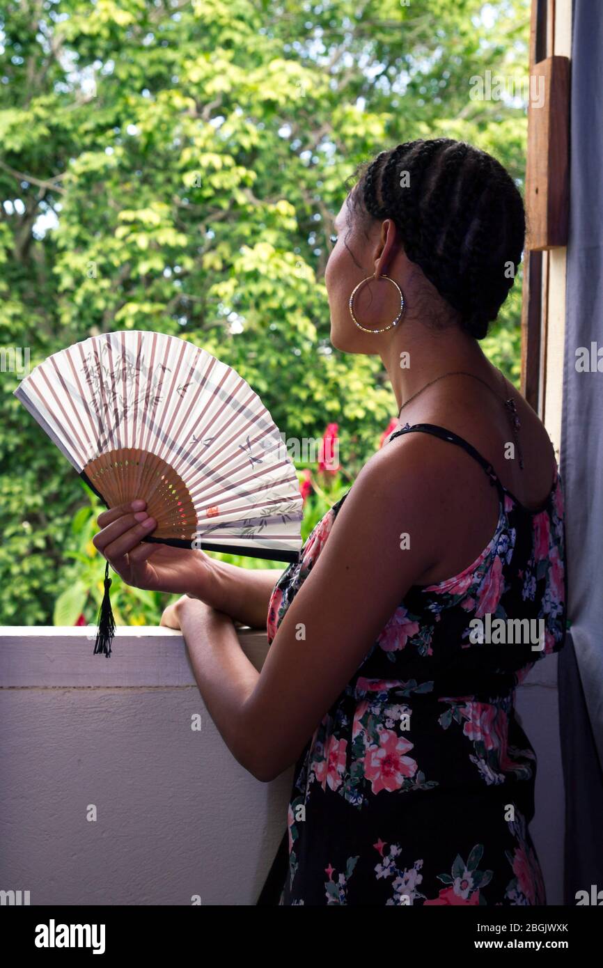 beautiful tanned young girl wearing long black dress and holding japanese fan leaning in a window inside a house with view of outside Stock Photo