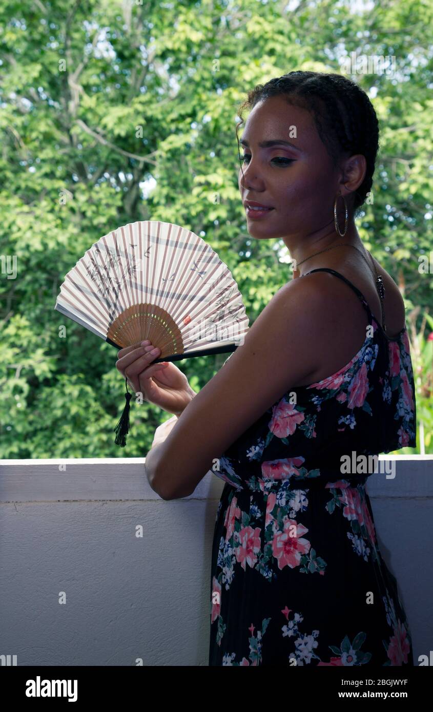 beautiful tanned young girl wearing long black dress and holding japanese fan leaning in a window inside a house with view of outside Stock Photo