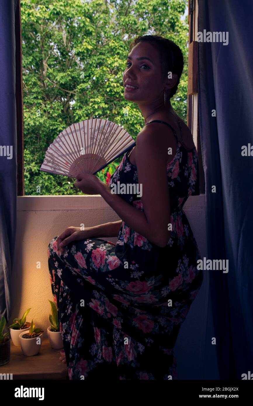 beautiful tanned young girl wearing long black dress and holding japanese fan leaning in a window inside a house with view of outside Stock Photo