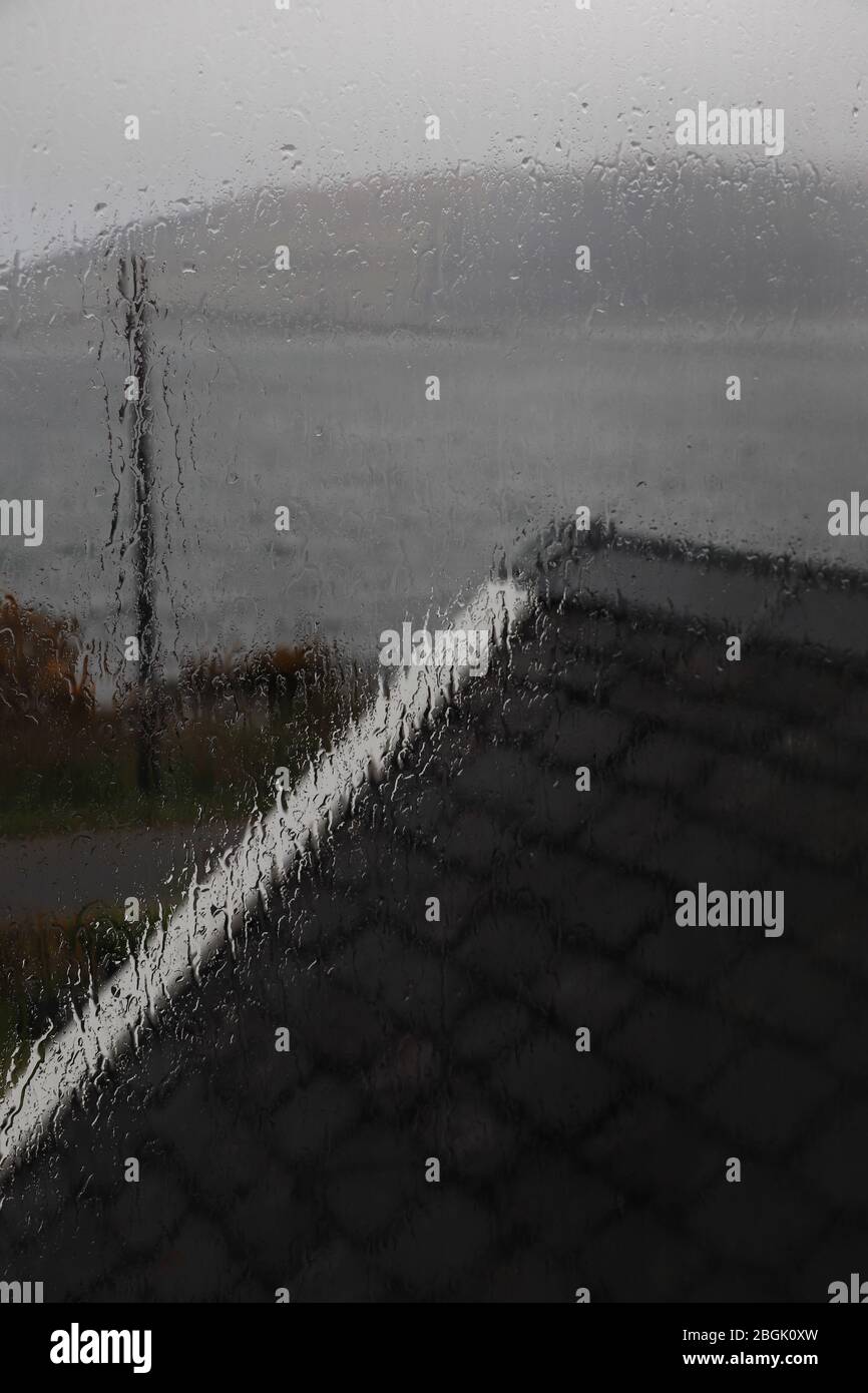 View out a rain splashed window, water running down, across a black slate roof with white trim to a loch + distant hill on a dark, dreary Scottish day Stock Photo