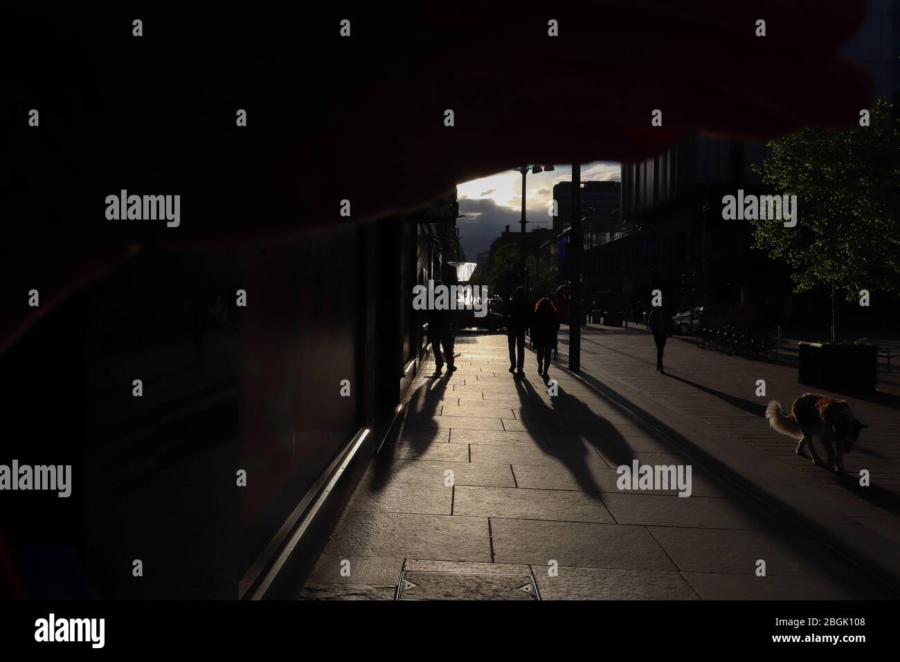 Concept 'Light at the end of the tunnel.' Several people in silhouette cast long shadows down a sidewalk in a backlit section of a street in Glasgow. Stock Photo