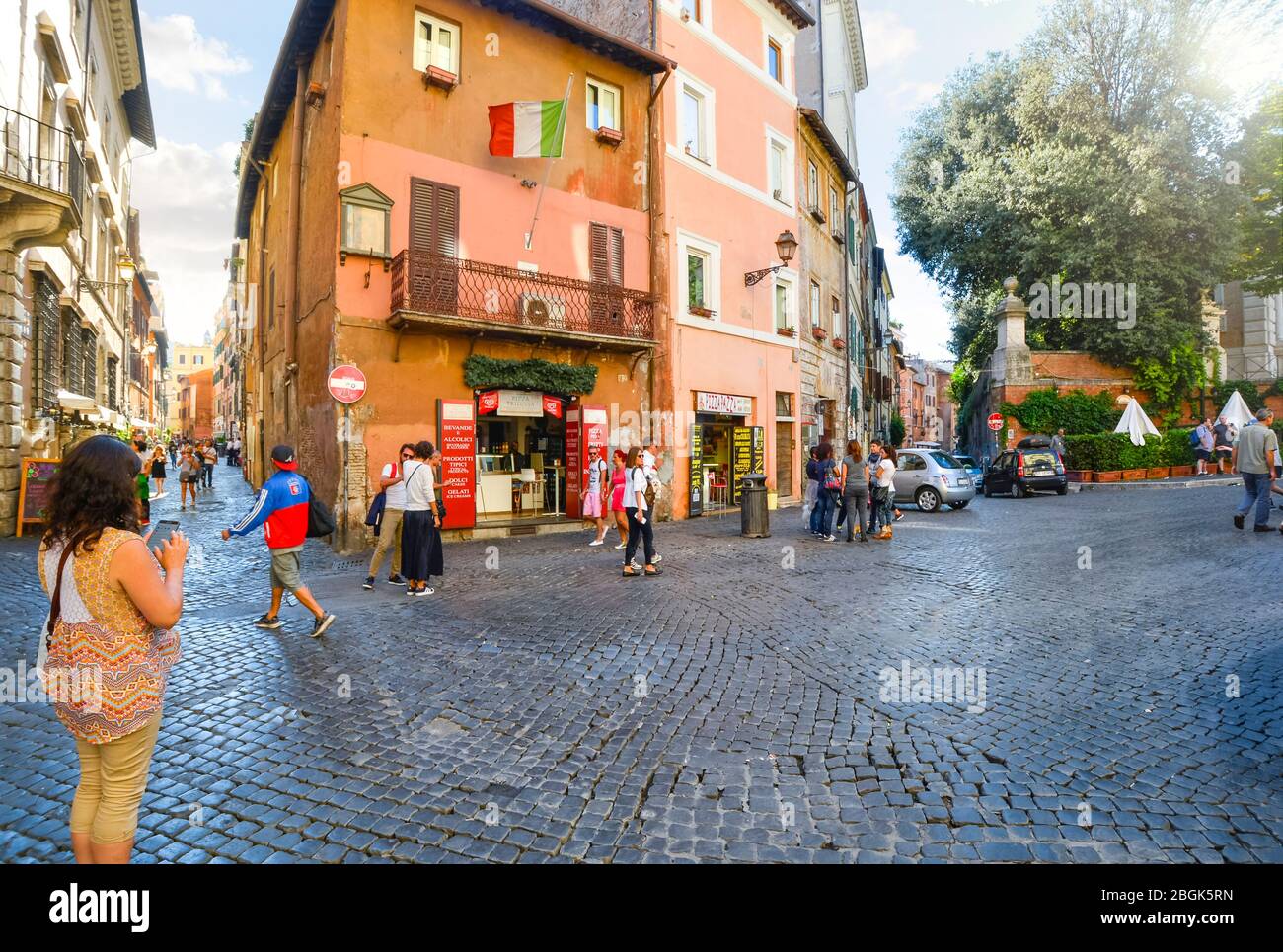 A female tourist uses her cell phone as other tourists walk by the shops and cafes in a small piazza in the Trastevere district of Rome, Italy. Stock Photo