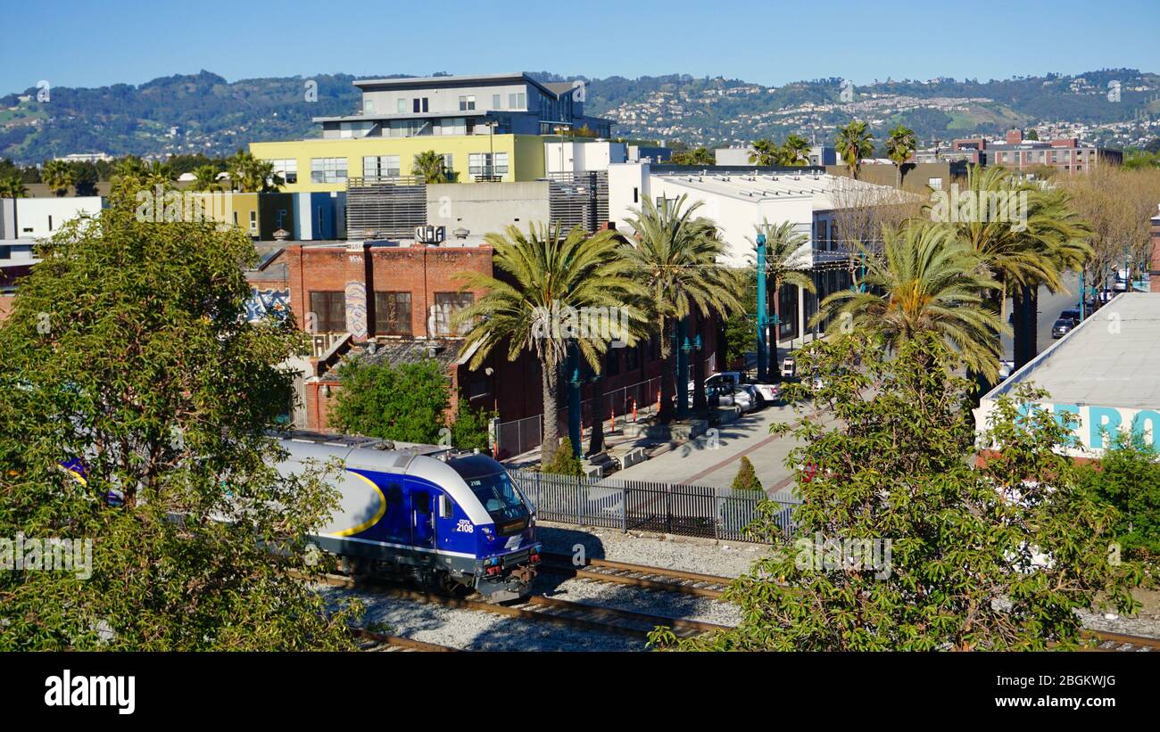 Amtrak passenger train passing through Emeryville. View of the Emeryville Warehouse Lofts and Oakland Hills in the background with palm trees. CA, USA Stock Photo