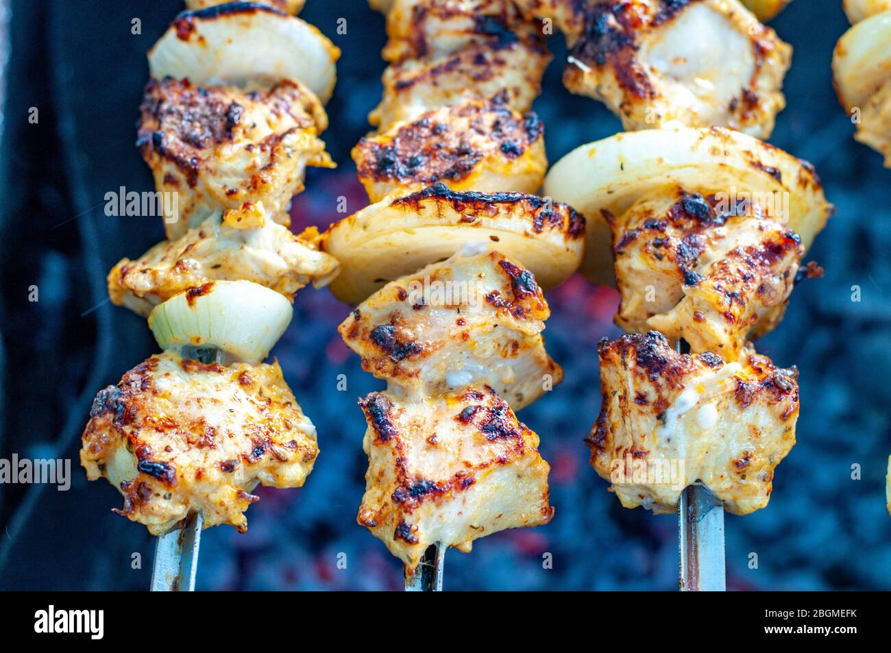 Roasted homemade chicken shish kabobs with fresh onions closeup atop smoldering coals in mangal. Grilled chicken meat skewers on blurred background. Stock Photo