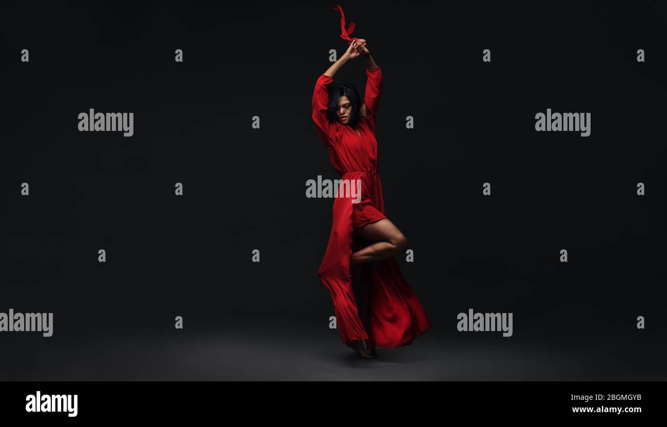 Full length of a young elegant woman dancing contemporary dance. Female in red dress performing strong dance moves over black background. Stock Photo