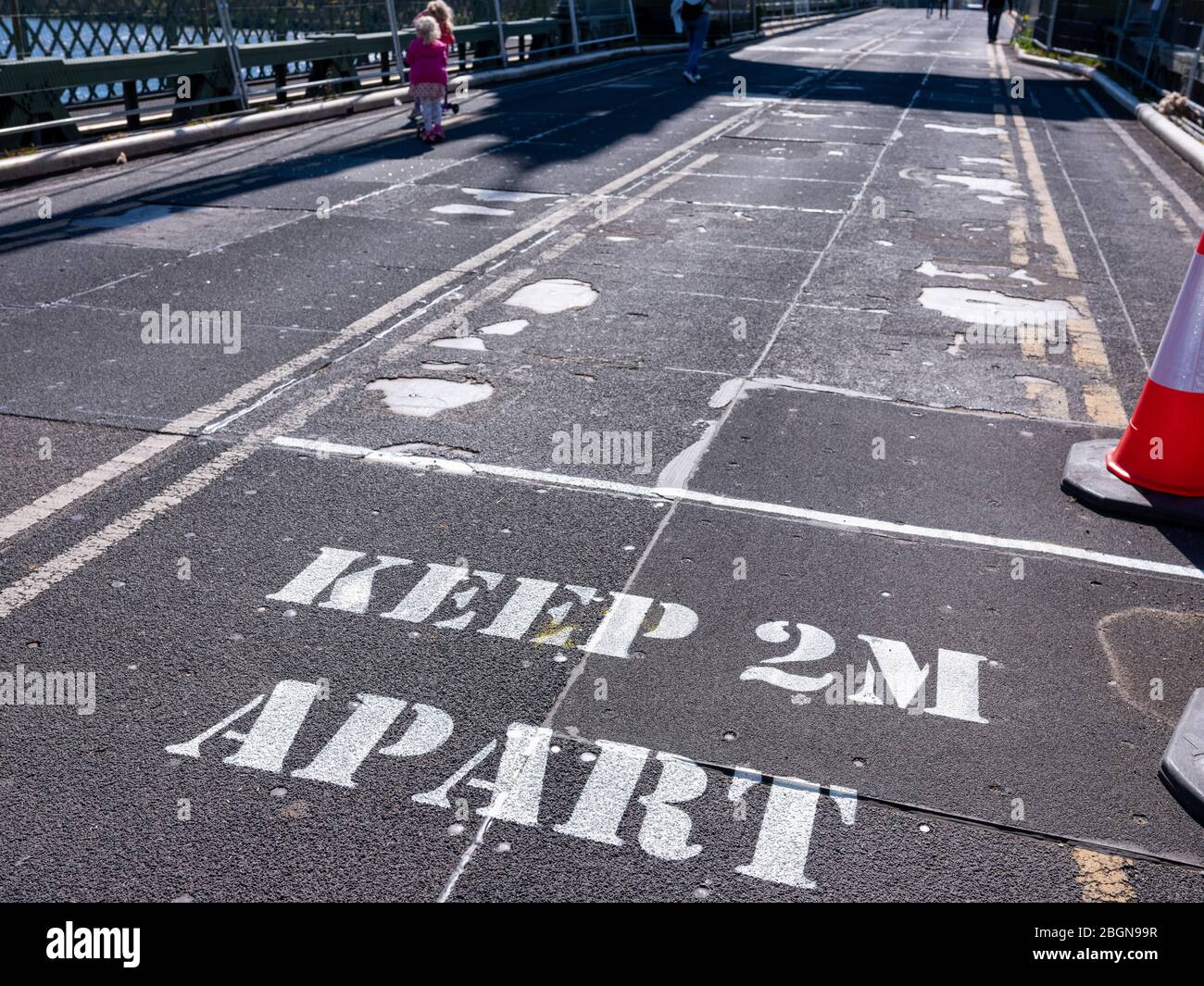 5th April 2020 A sign painted on the road surface warns pedestrians to keep their distance in order to prevent the Covid-19 virus spreading. Two young Stock Photo