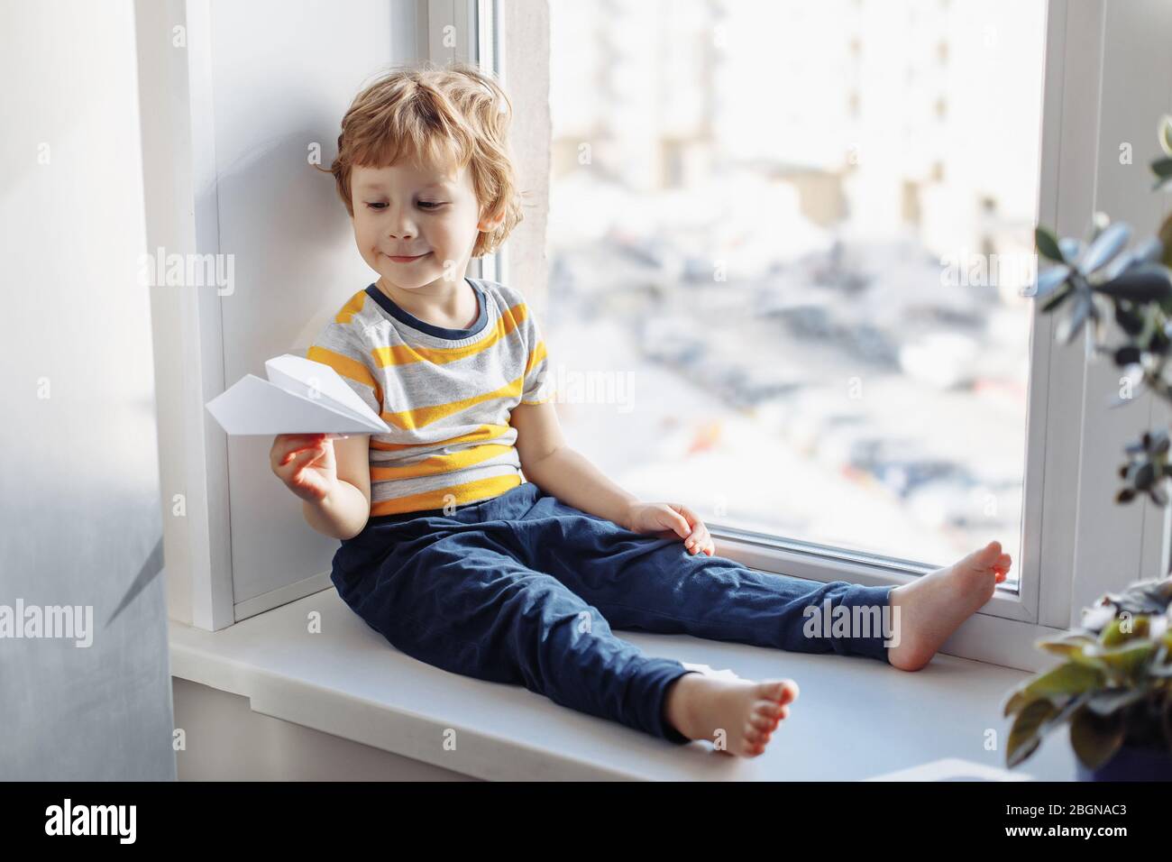 Child boy sitting near window with paper plane and looking. Stay home ...