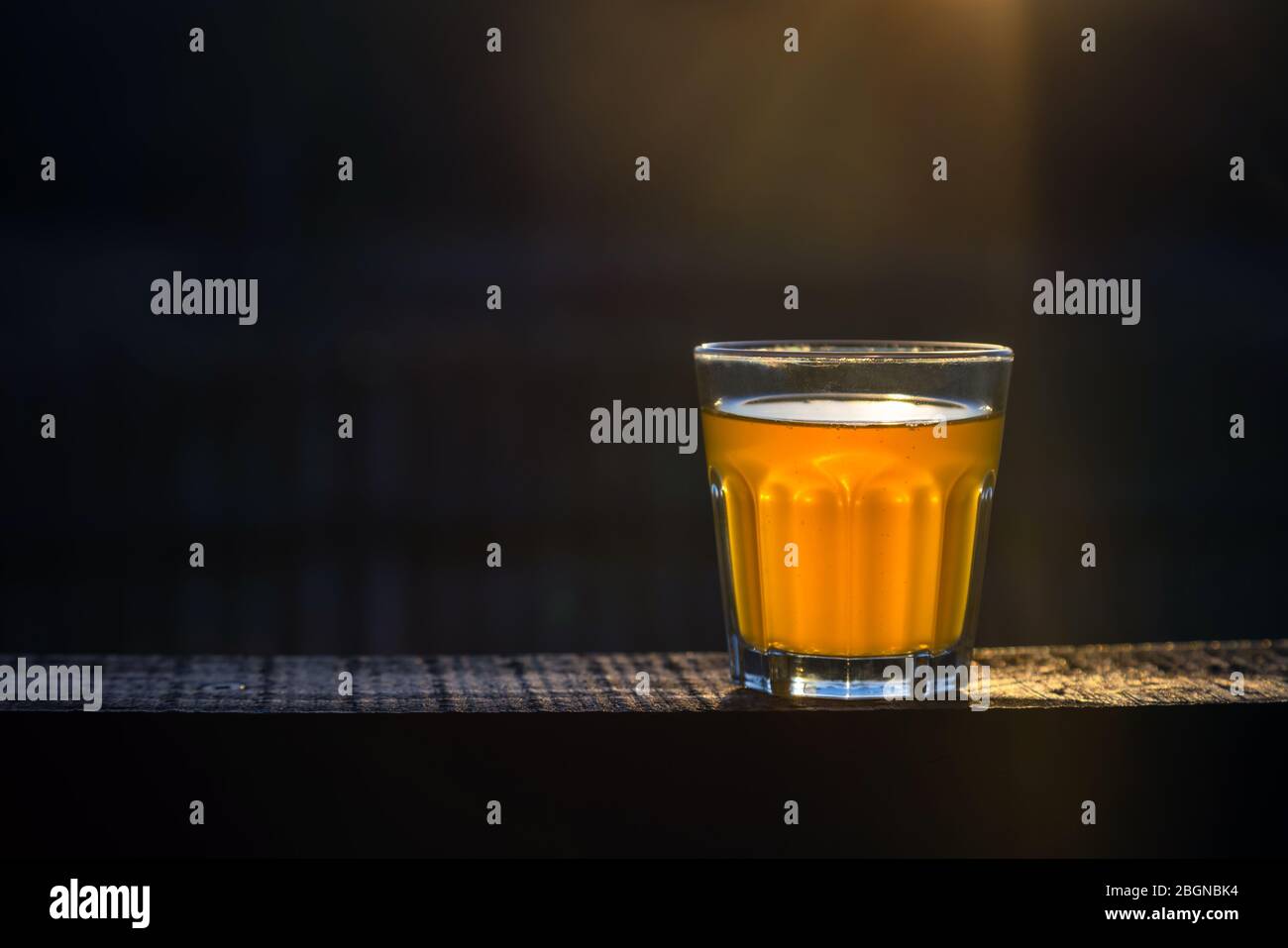 Small glass of craft beer with no foam on a wooden plank outside with space for text. Golden sample of alcohol beverage on dark background. Nobody. Stock Photo