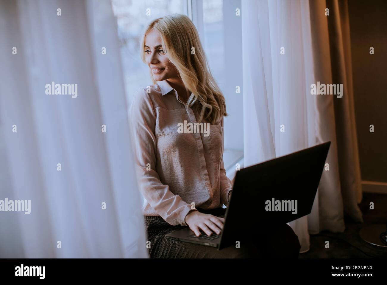 Pretty young woman working on laptop in living room on the window sill ...