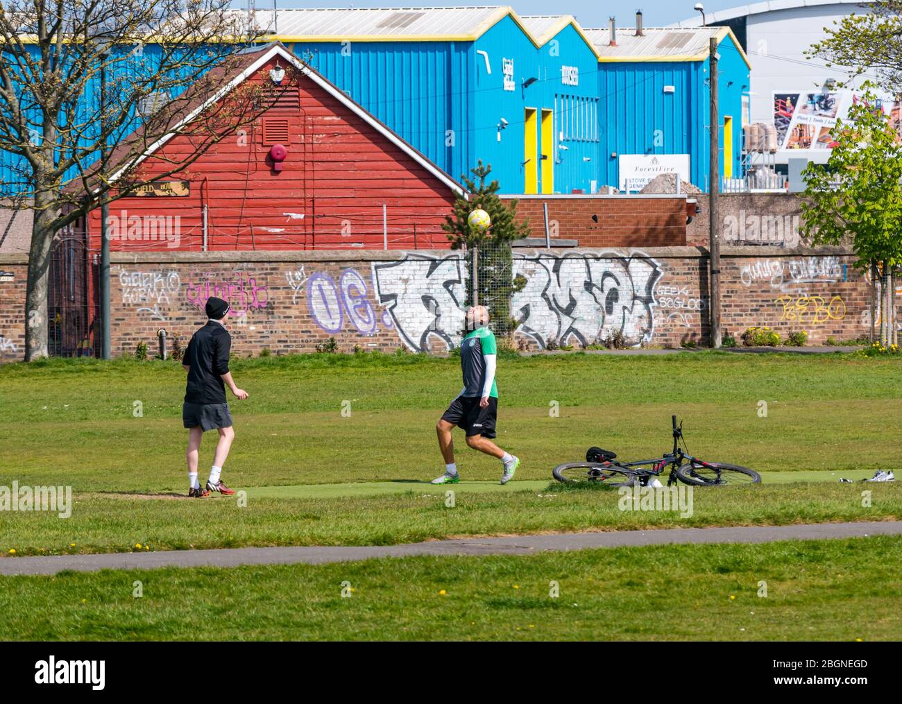 Leith, Edinburgh, Scotland, United Kingdom. 22nd April 2020. Covid-19 Lockdown: people exercise on Leith Links in Spring sunshine. Two men kick a football back and forth between each other in their daily exercise during the Coronavirus pandemic Stock Photo