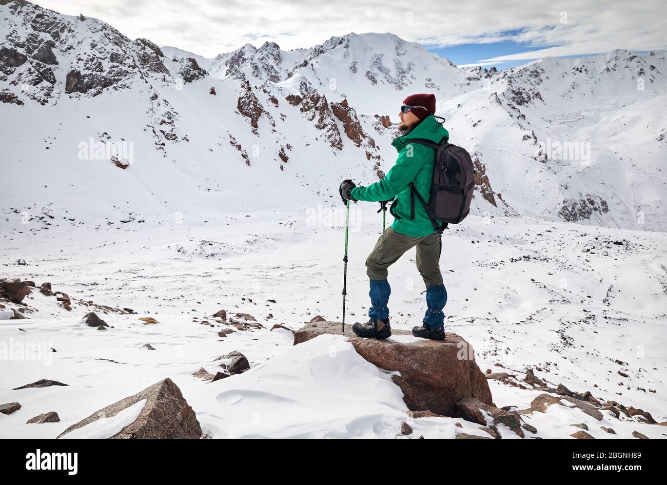Hiker with trekking poles and backpack standing on the rock at snowy mountains background Stock Photo