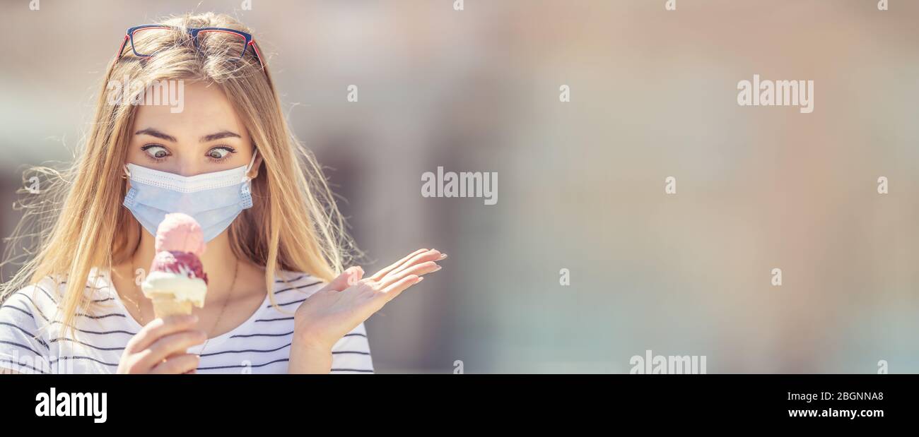 Beautiful girl wearing a face mask looking down at an ice cream in her hand with a cross-eyed look and deperate hand gesture. Stock Photo