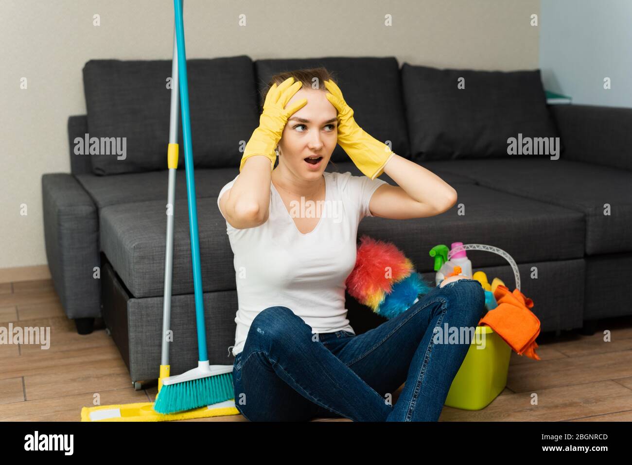 A beautiful woman next to cleaning products and equipment is sitting on the floor and screaming, holding on to her head. A young girl does the cleaning in the house. Stock Photo