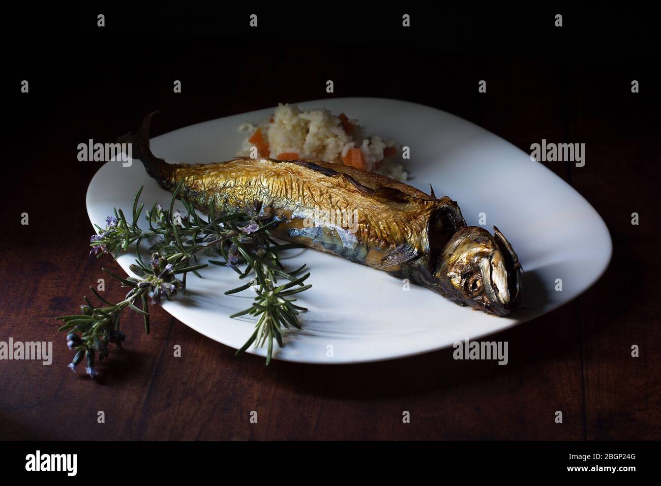 Oven-roasted mackerel, served on a white plate, a small portion of cooked rice with carrot pieces, and a few flowering rosemary twigs as a decoration. Stock Photo
