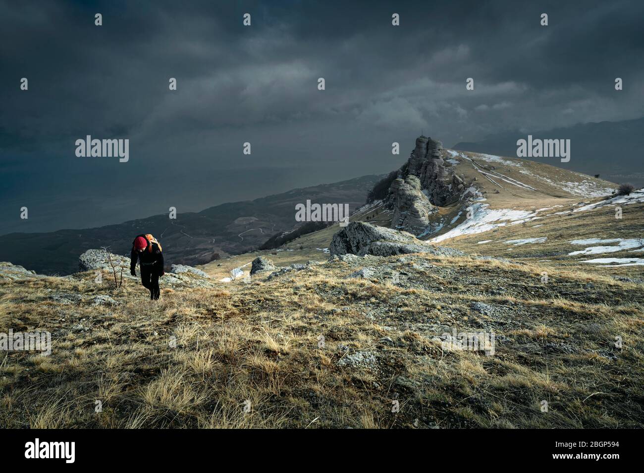 Female hiker with an orange backpack goes up the mountain. Overcast day. Snow on the rocks. Dry grass. Horizontal image for winter stories Stock Photo