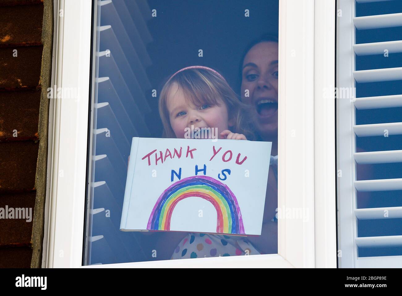Brighton UK 22nd April 2020 - A young girl and her mother stick a Thank You NHS rainbow painting and message to their Woodingdean house window near Brighton during lockdown in the Coronavirus COVID-19 pandemic crisis  . Credit: Simon Dack / Alamy Live News Stock Photo