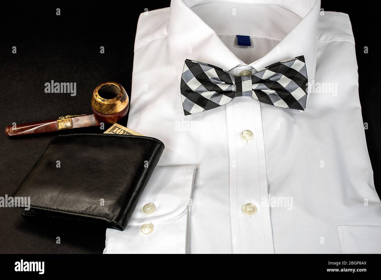black and white checkered bow tie on folded white dress shirt with wallet and old smoking pipe on black Stock Photo