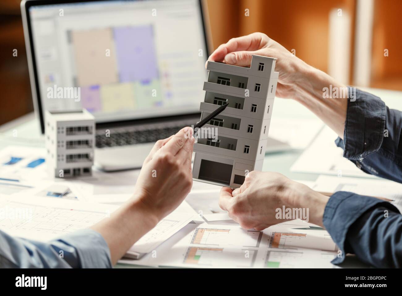 Two architects holding and discussing model of a house and architectural plans in a studio with blueprints and laptop on a table. Architectural desigh Stock Photo