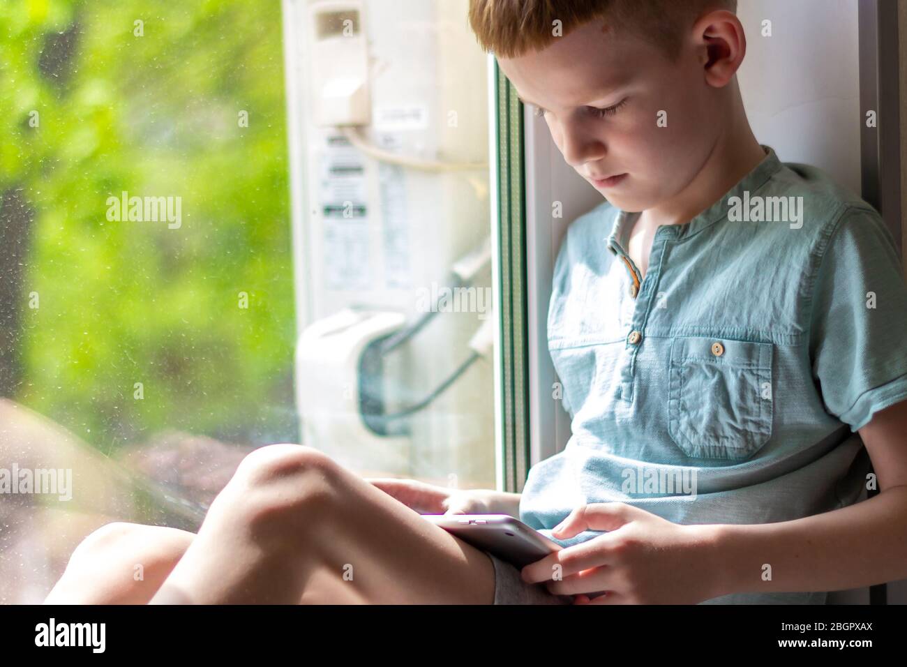 Young boy sitting on windowsill with tablet and play the games. Stay at home during quarantine. Stock Photo