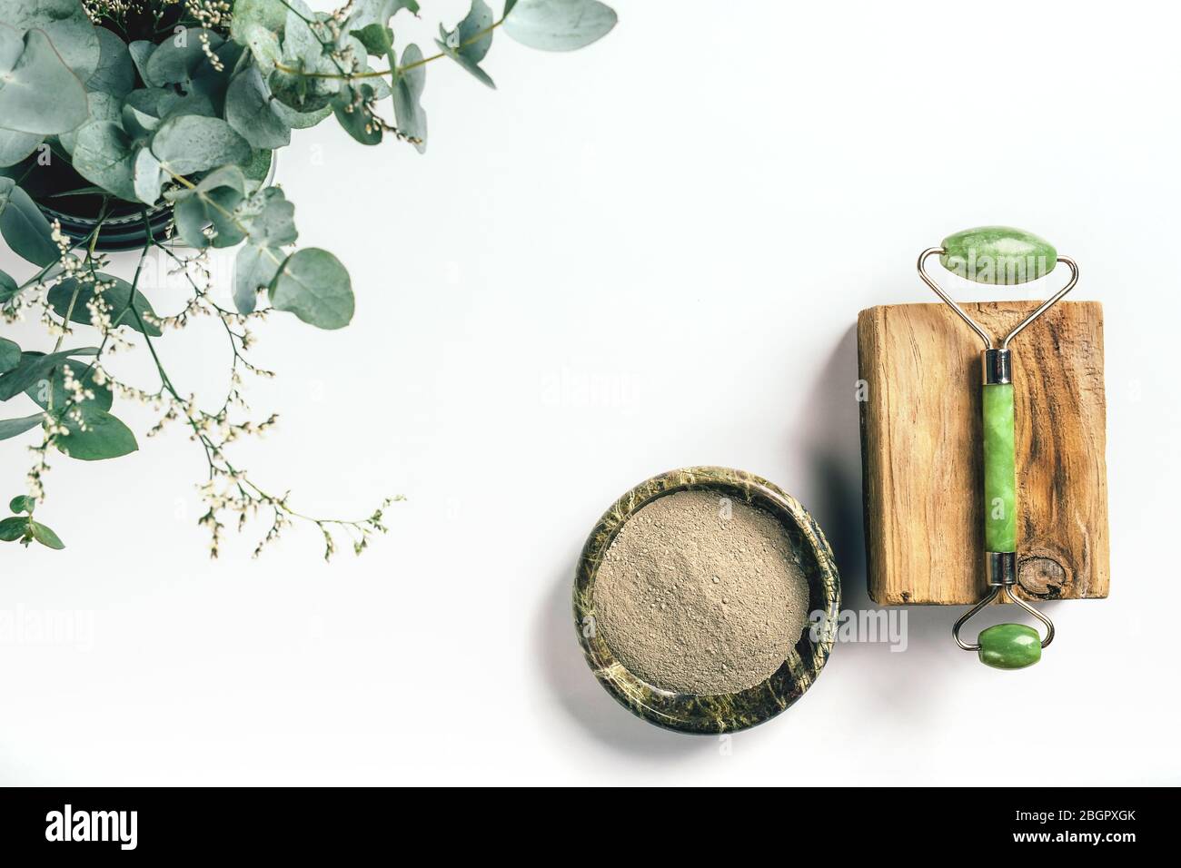 Facial greenstone roller on wooden bar and clay powder in bowl with eucalyptus and herbs in the corner on white paper background with copy space. Health and beauty concept. Stock Photo