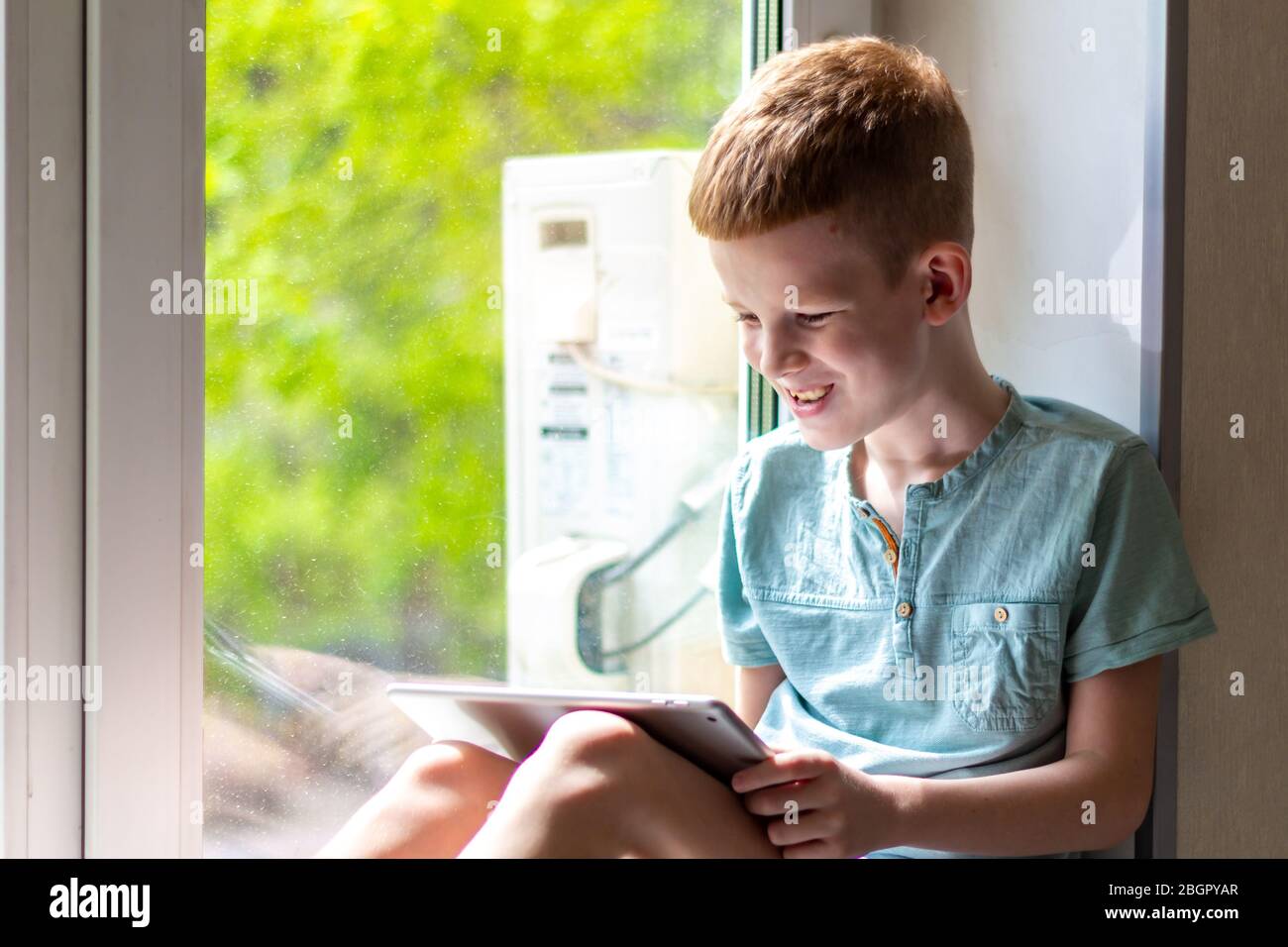 Young boy sitting on windowsill with tablet and play the games. Stay at home during quarantine. Stock Photo